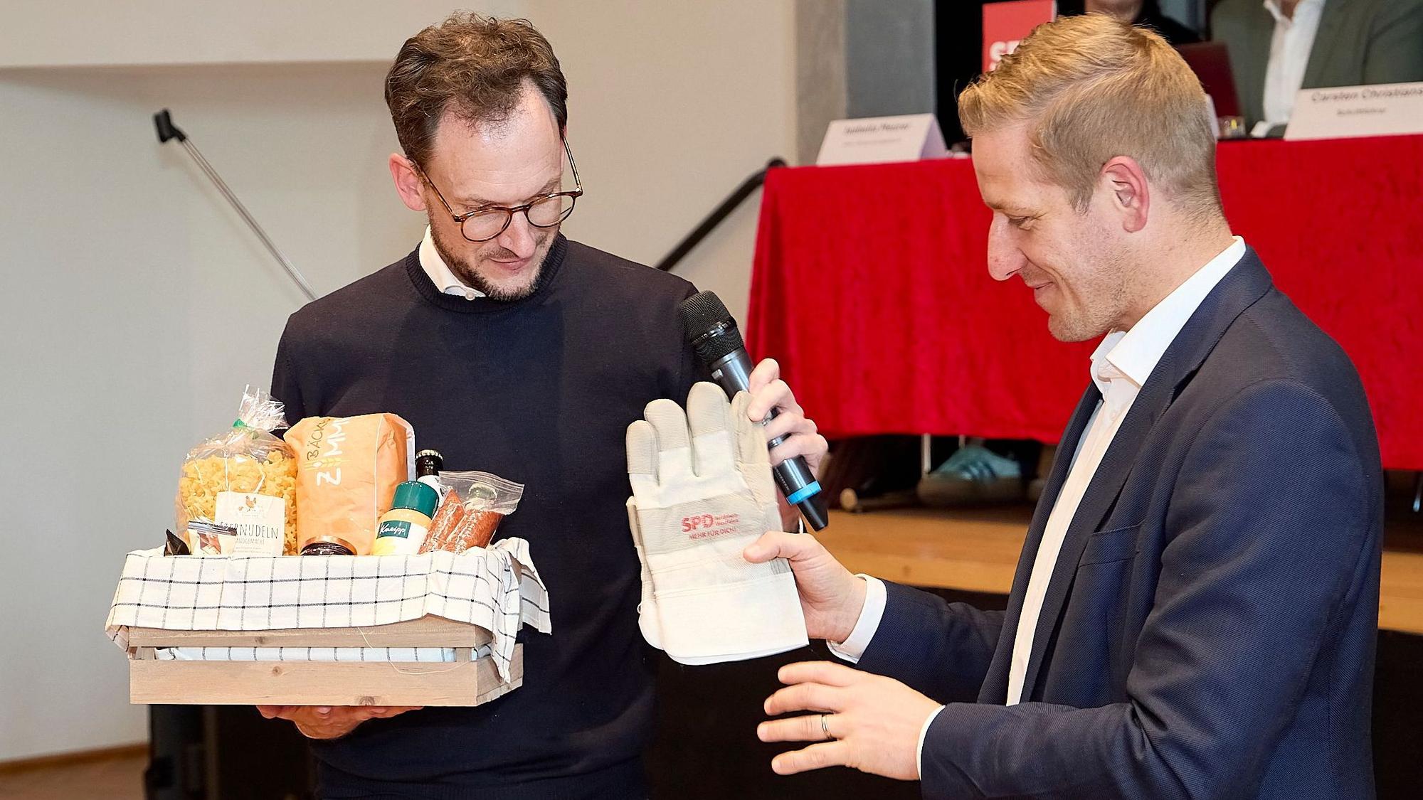 Das Bild zeigt, wie Waasem Arbeitshandschuhe mit der Aufschrift SPD an Markus Ramers übergibt. In der anderen Hand hält Waasem einen Fresskorb