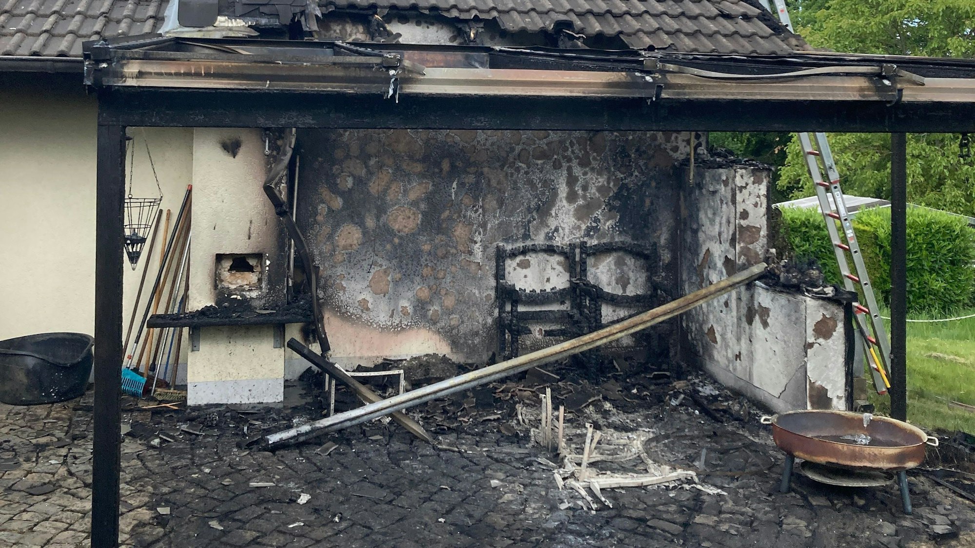 An einer verkohlten Garagenwand stehen verbrannte Stühle.
