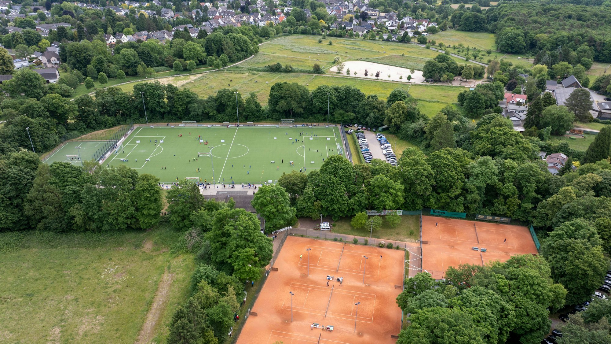 Eingekeilt zwischen Tennisplätzen und dem Kunstrasenplatz des Sportvereins Adler Dellbrück liegt einer kleinen Senke ein alter Sportplatz (links unten), der von Bäumen und Büschen umrandet und mit Gras bewachsen ist. (Luftaufnahme mit Drohne.)