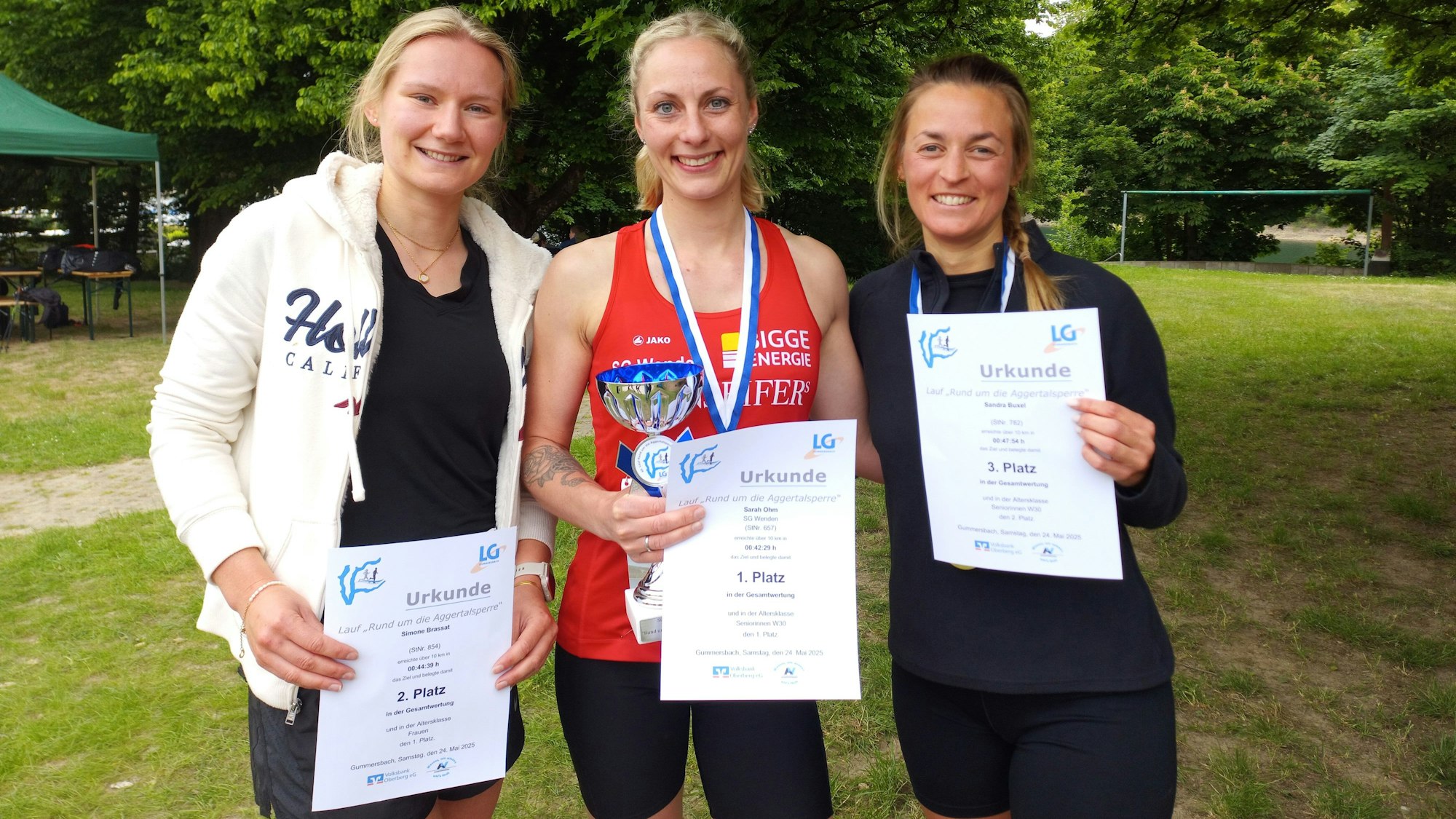 Zu sehen sind drei Frauen, die Urkunden und einen Pokal in den Händen halten.