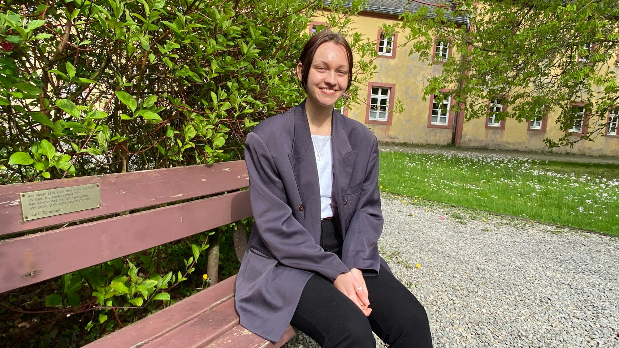 Myra Abel sitzt auf einer Bank, im Hintergrund sieht man Gebäude des Klosters Steinfeld.
