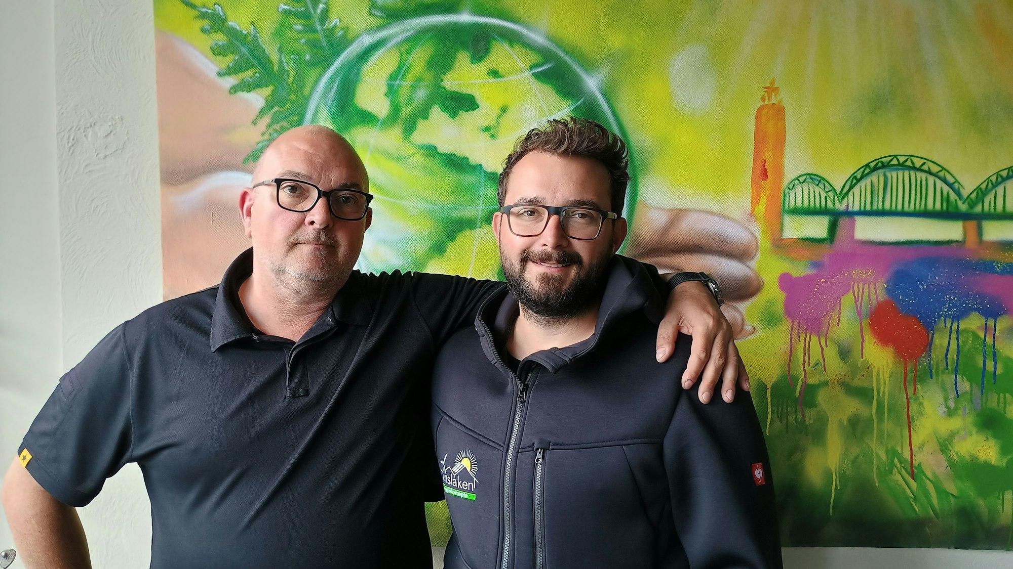 Oliver Dinslaken hat den Arm um seinen Sohn Max gelegt. Hinter ihnen ist ein Graffito, das einen grünen Globus und die Kölner Hohenzollernbrücke zeigt.