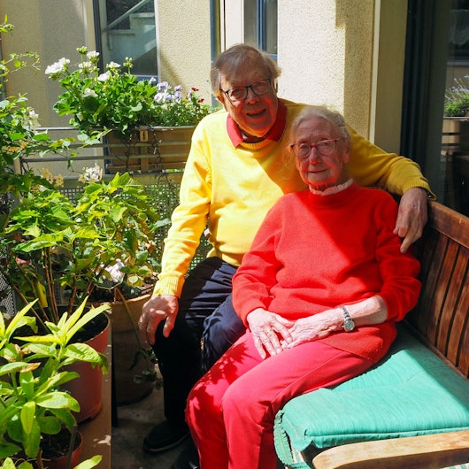 Auf einem Balkon mit vielen blühenden Pflanzen sitzen ein älterer Mann und eine ältere Dame auf einer Bank. Er hat seinen Arm um sie gelegt.