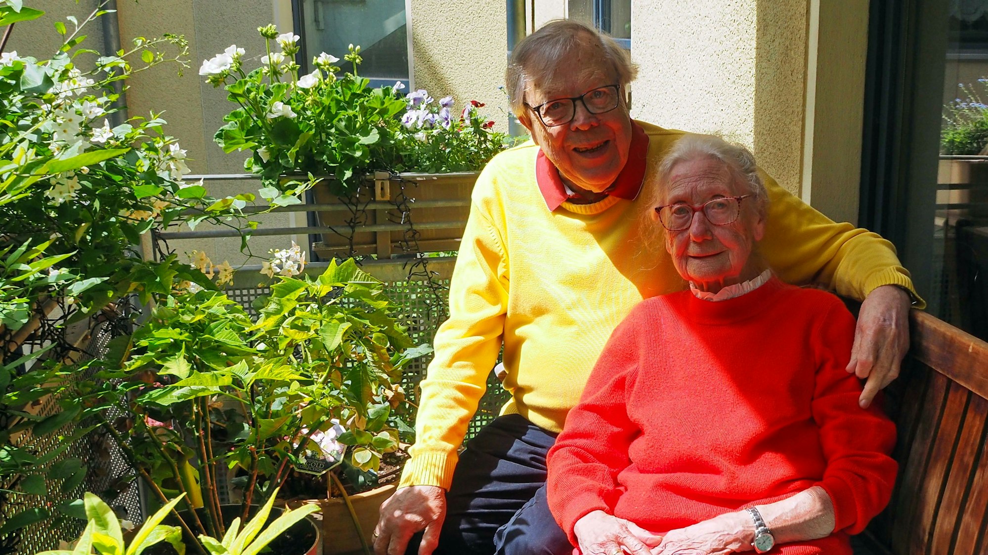 Auf einem Balkon mit vielen blühenden Pflanzen sitzen ein älterer Mann und eine ältere Dame auf einer Bank. Er hat seinen Arm um sie gelegt.