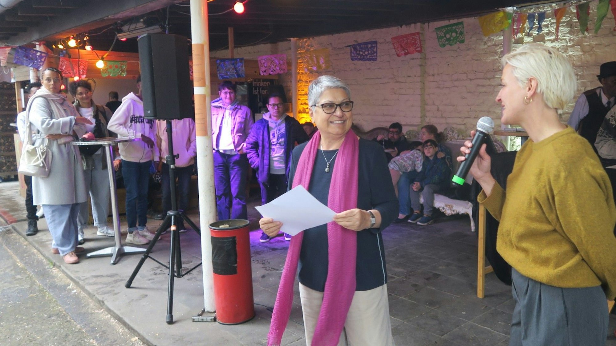 Zwei Frauen stehen unter einem Vordach, eine hält einen Zettel, die andere ein Mikrofon in der Hand.
