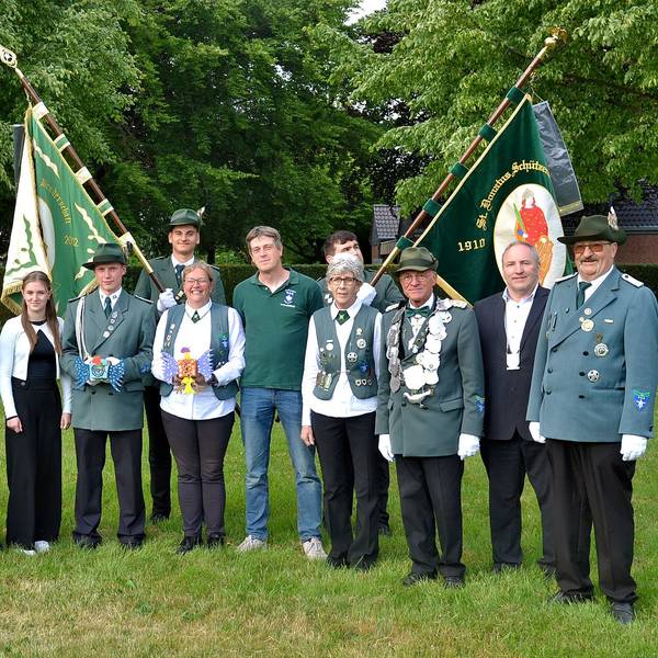 Eine Gruppe Menschen steht auf einer Wiese, manche tragen eine grün Uniform, im Hintergrund sind zwei Fahnen zu erkennen.