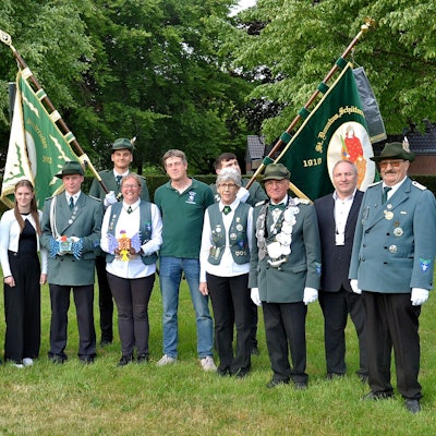 Eine Gruppe Menschen steht auf einer Wiese, manche tragen eine grün Uniform, im Hintergrund sind zwei Fahnen zu erkennen.