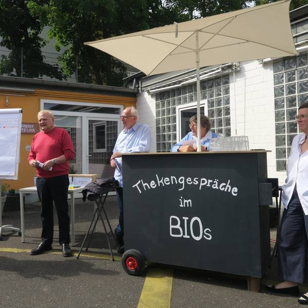 Drei Männer und zwei Frauen stehen in einem Innenhof. Sie haben eine mobile Theke dabei, auf der steht: Thekengespräche im Bios.