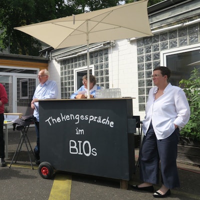 Drei Männer und zwei Frauen stehen in einem Innenhof. Sie haben eine mobile Theke dabei, auf der steht: Thekengespräche im Bios.