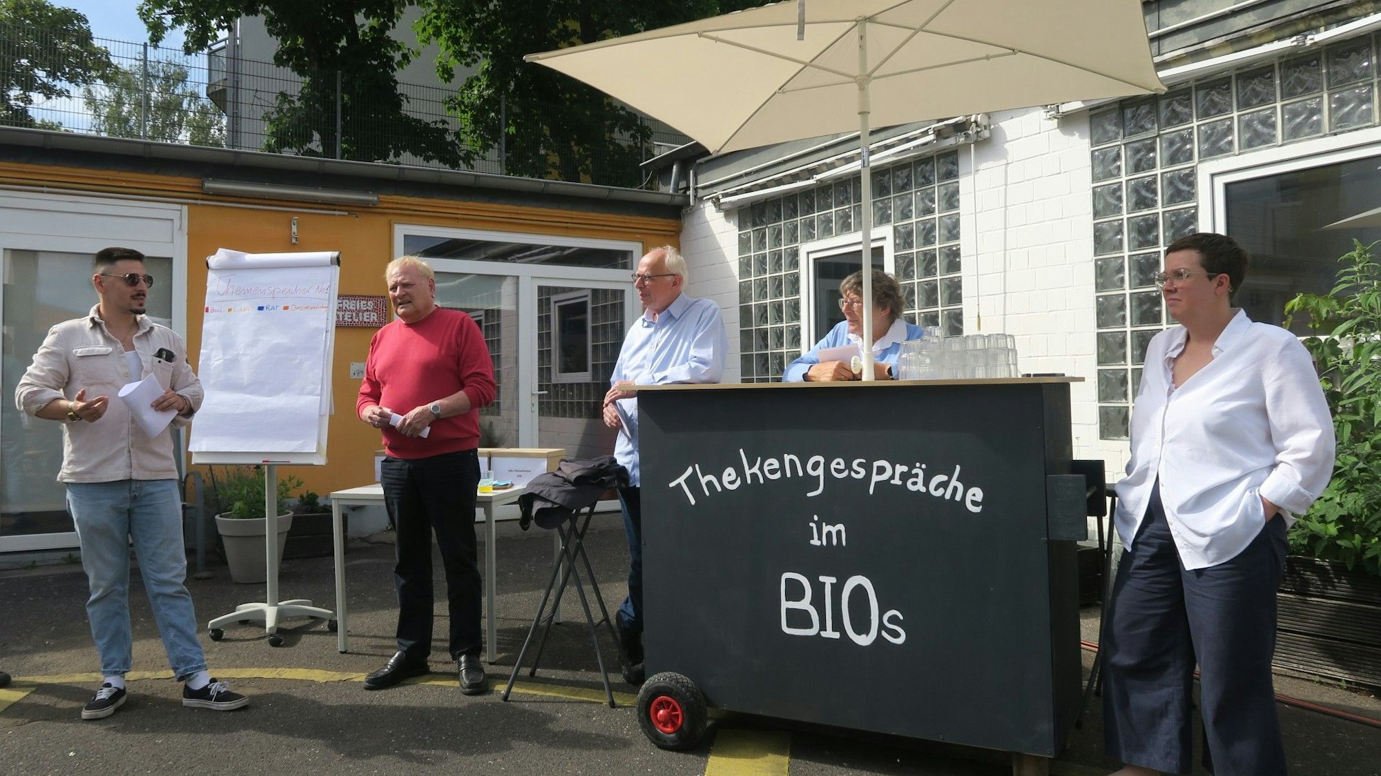 Drei Männer und zwei Frauen stehen in einem Innenhof. Sie haben eine mobile Theke dabei, auf der steht: Thekengespräche im Bios.