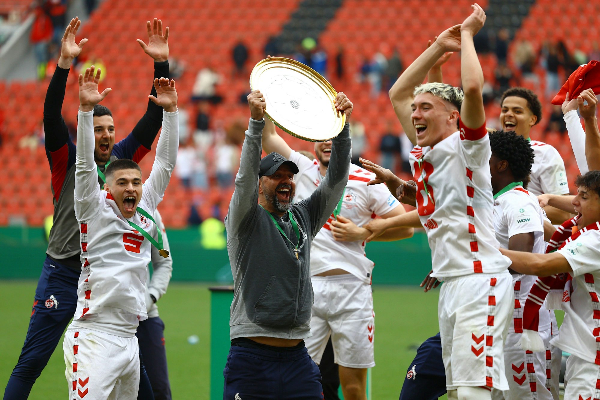 Ein Fußballtrainer reckt eine Meisterschale in die Höhe. Seine Spieler freuen sich.