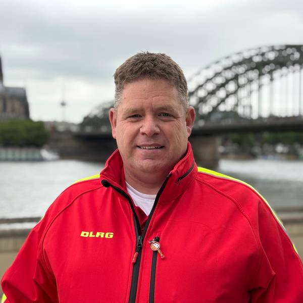 Frank Zantis steht in einer knallroten Weste der DLRG am Rheinufer. Kölner Dom und Hohenzollernbrücke im Hintergrund sind unscharf gestellt.