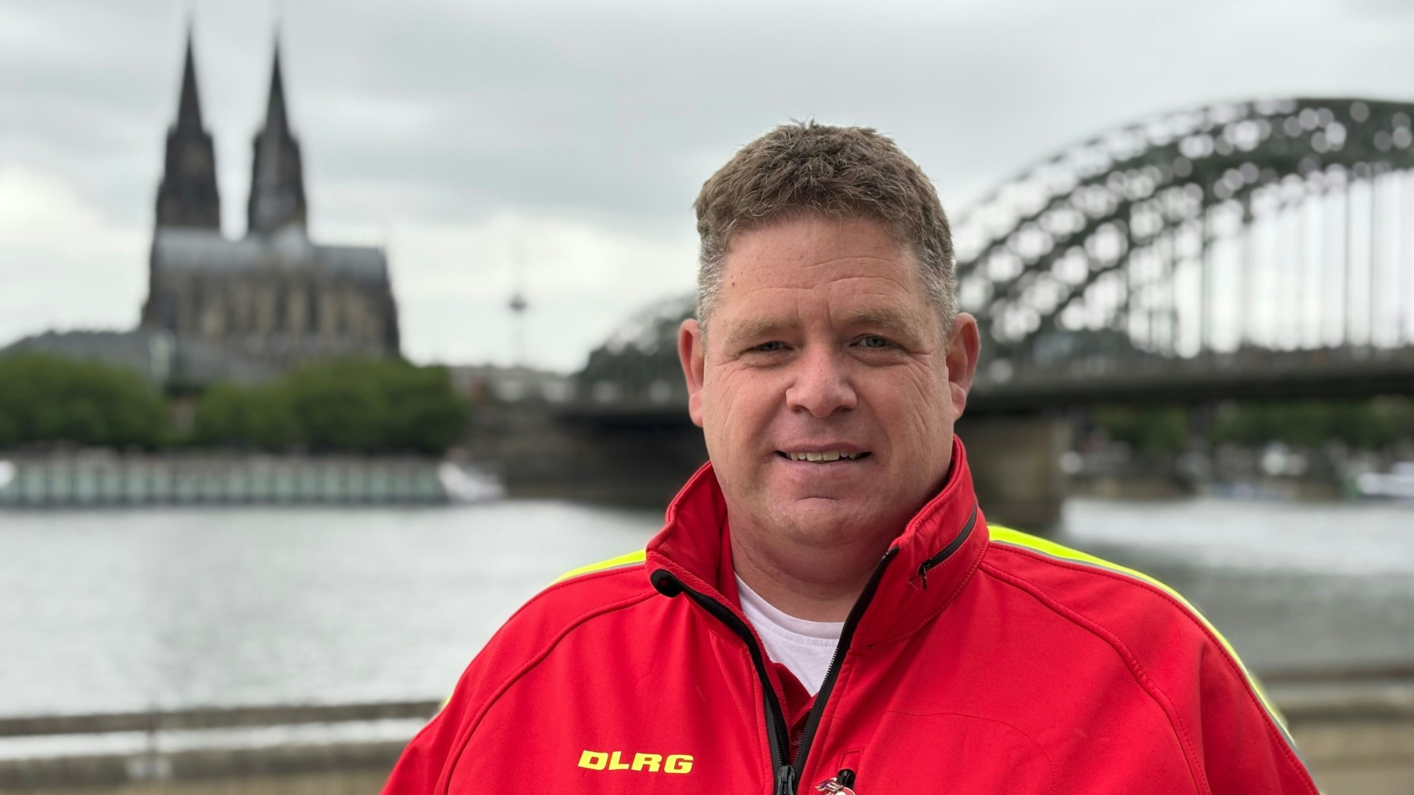 Frank Zantis steht in einer knallroten Weste der DLRG am Rheinufer. Kölner Dom und Hohenzollernbrücke im Hintergrund sind unscharf gestellt.