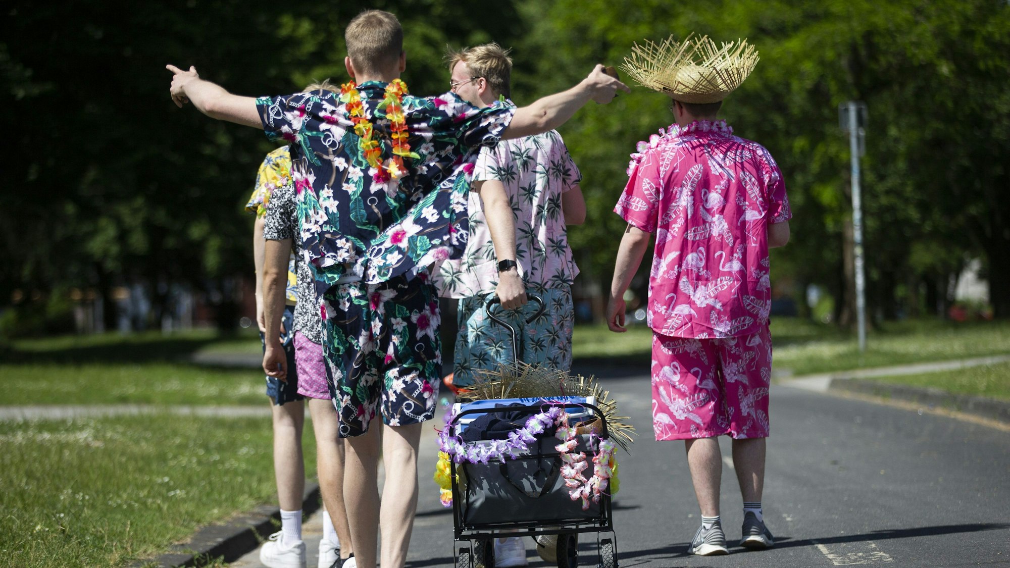 Junge Männer in Outfits mit floralen Mustern gekleidet, ziehen einen geschmückten Bollerwagen mit Getränken am Aachener Weiher in Köln hinter sich her. (Archivbild)