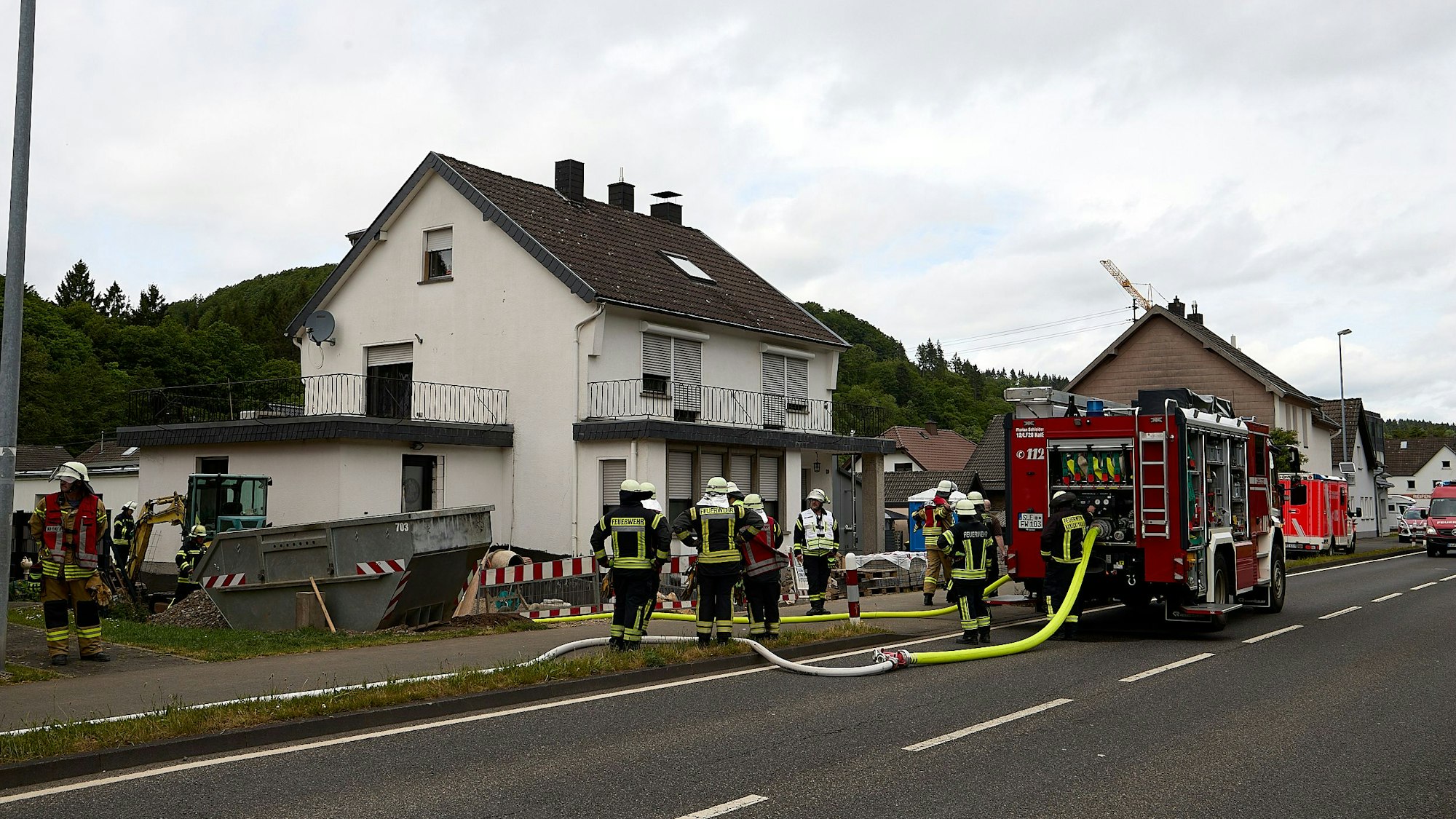 Mehrere Feuerwehrleute stehen vor einem weißen Haus. Auf der Straße steht ein Feuerwehrfahrzeug, im Hintergrund ist ein Rettungswagen zu sehen.