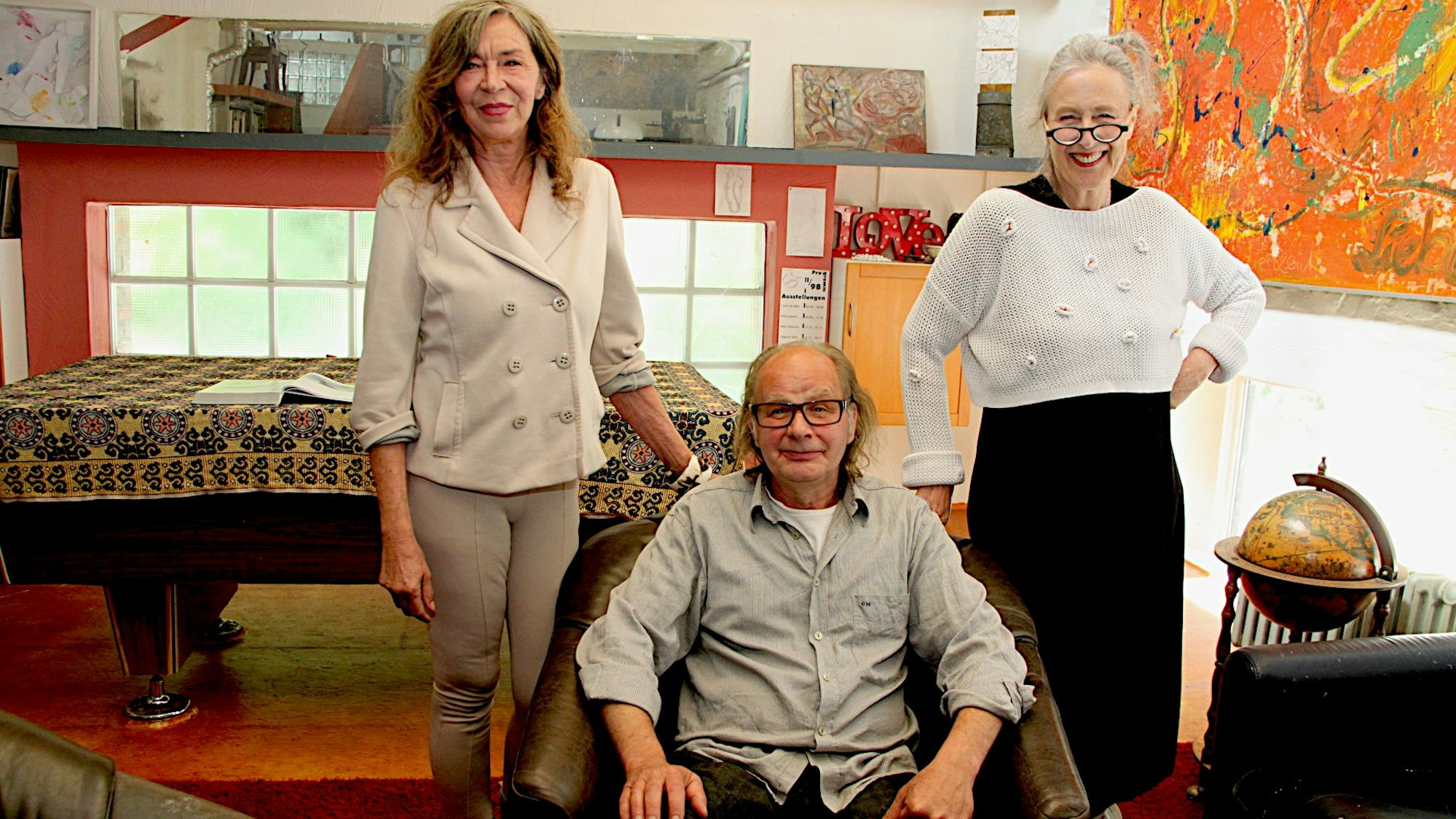 Peter Sußner sitzt in einem Sessel, er trägt dunkle Hose und hellgraues Hemd. Links und rechts neben dem Sessel stehen Carin Conscience und Monika Ertl-Berns.