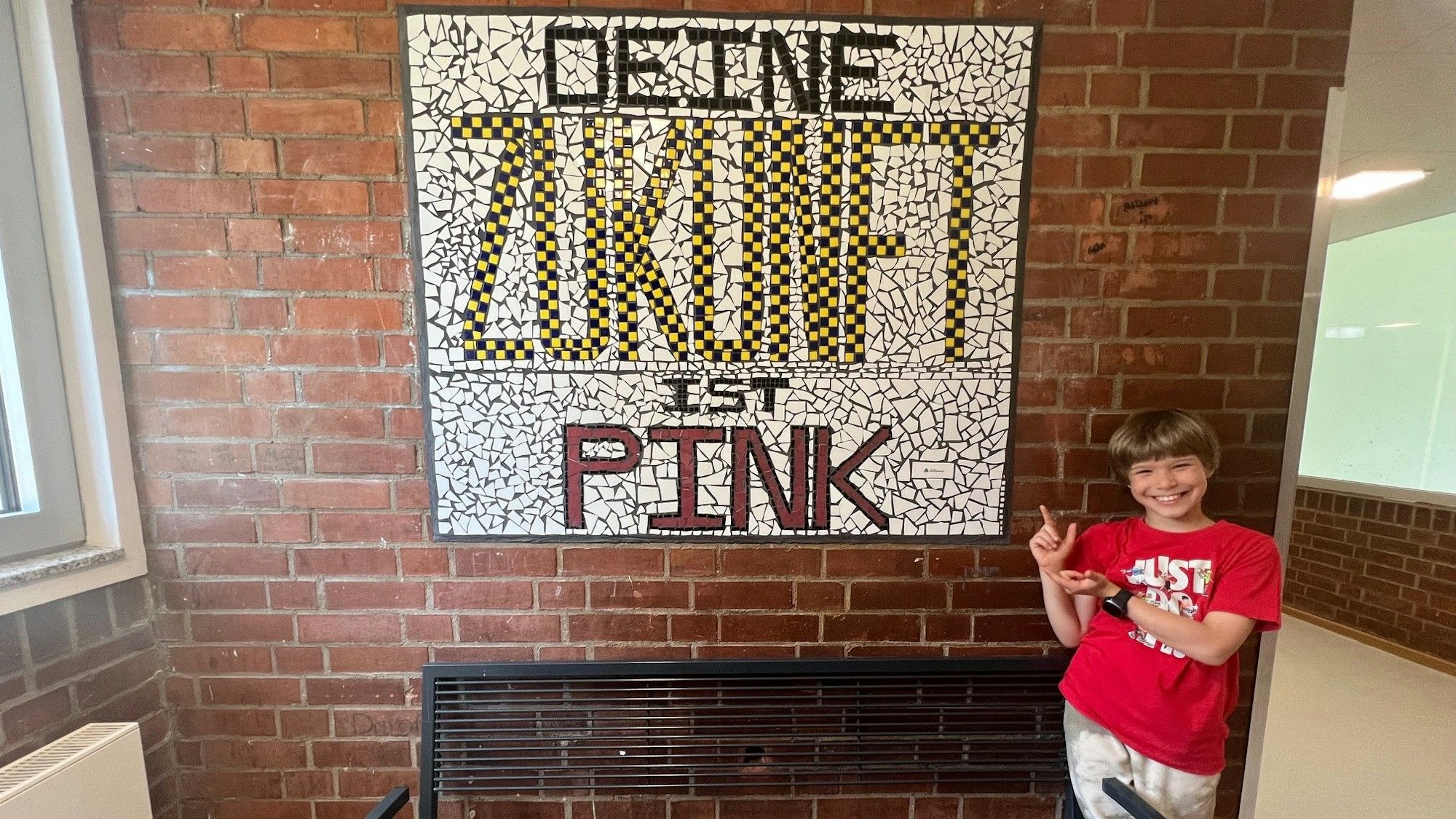 Ein Junge steht vor einem großen Mosaik, auf dem „Deine Zukunft ist pink“ steht, es hängt an einer Wand der Jugendeinrichtung OT Ohmstraße.