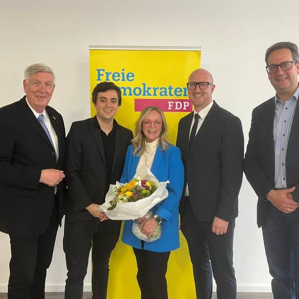 Die Herren haben die Kandidatin, die einen Blumenstrauß in den Händen hält, in ihre Mitte genommen. Dahinter steht ein Aufsteller mit der Aufschrift Freie Demokraten FDP.