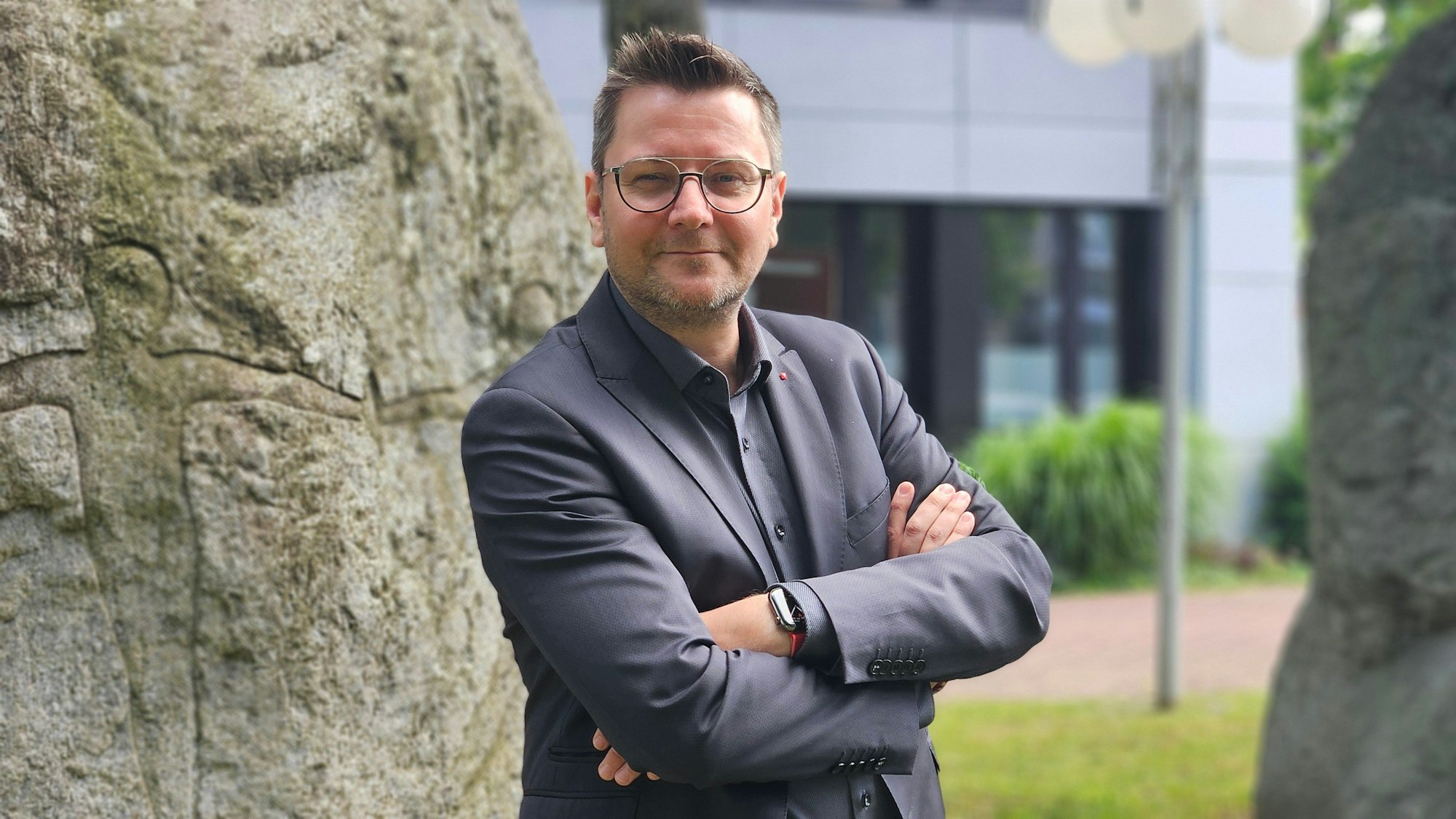 Auf dem Foto ist ein Mann mit Brille zu sehen. Er steht mit verschränkten Armen vor dem Baum.