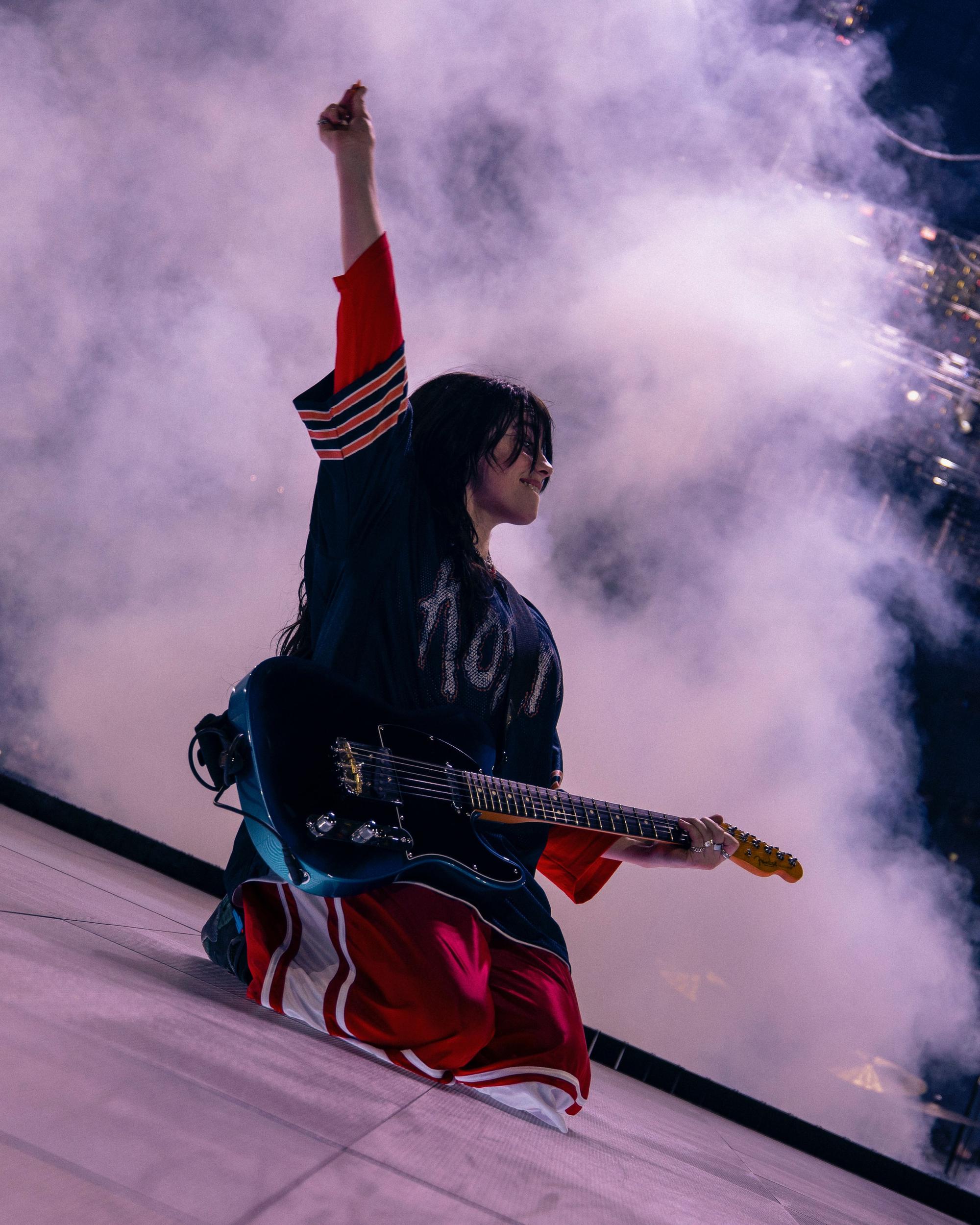 Billie Eilish kniet mit einer E-Gitarre auf der Bühne in der Lanxess-Arena. Den linken Arm hat sie hochgestreckt, in den Fingern hält sie ein Plektrum.
