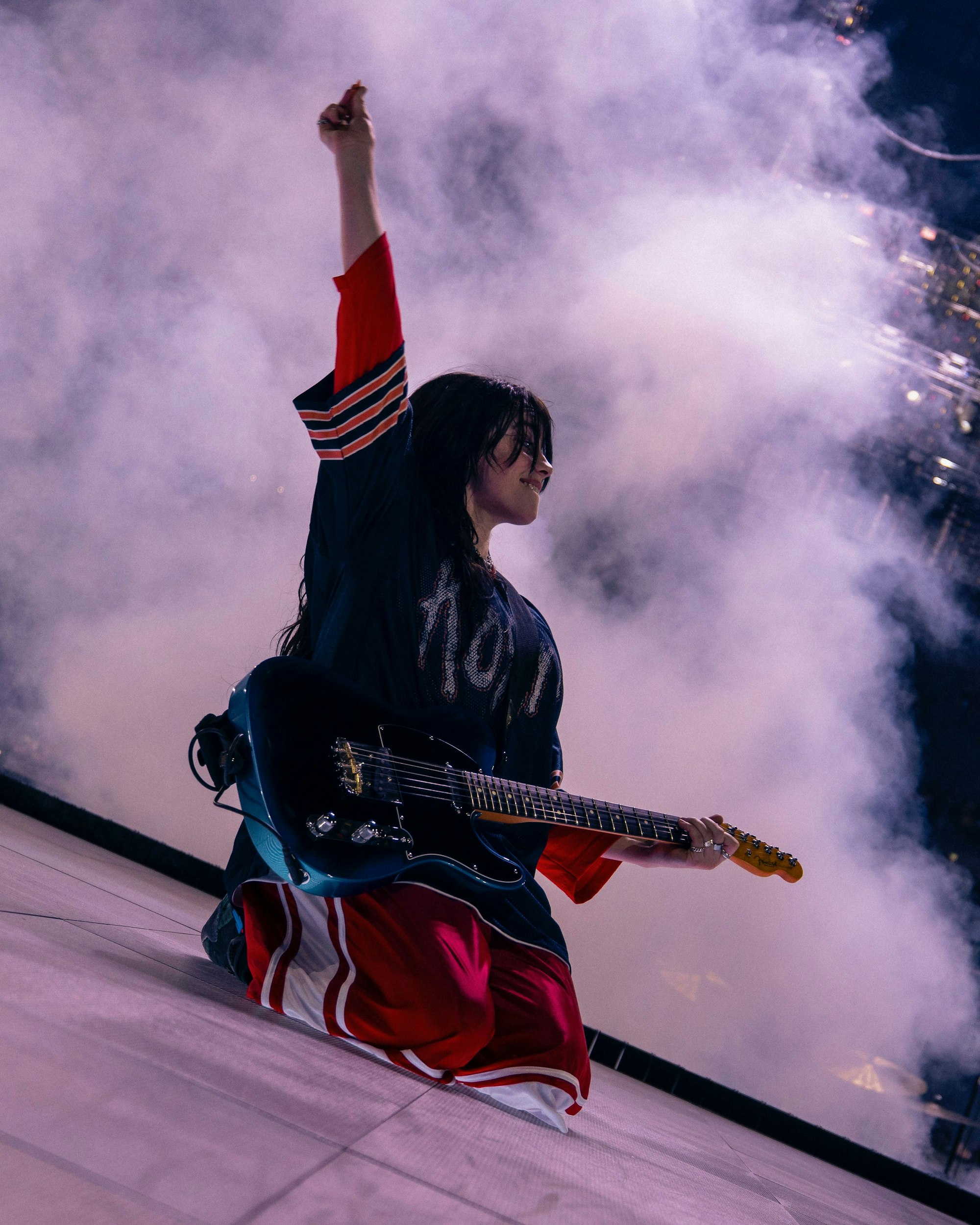 Billie Eilish kniet mit einer E-Gitarre auf der Bühne in der Lanxess-Arena. Den linken Arm hat sie hochgestreckt, in den Fingern hält sie ein Plektrum.