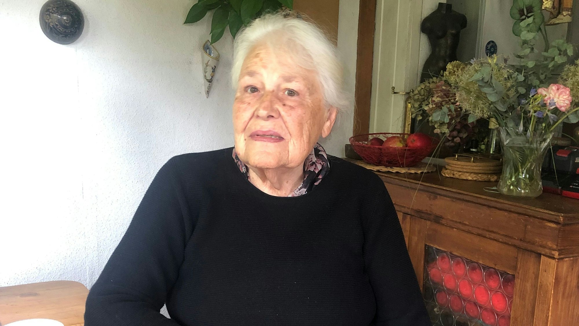 Eine ältere Frau sitzt an einem Esstisch und blickt traurig in die Kamera.