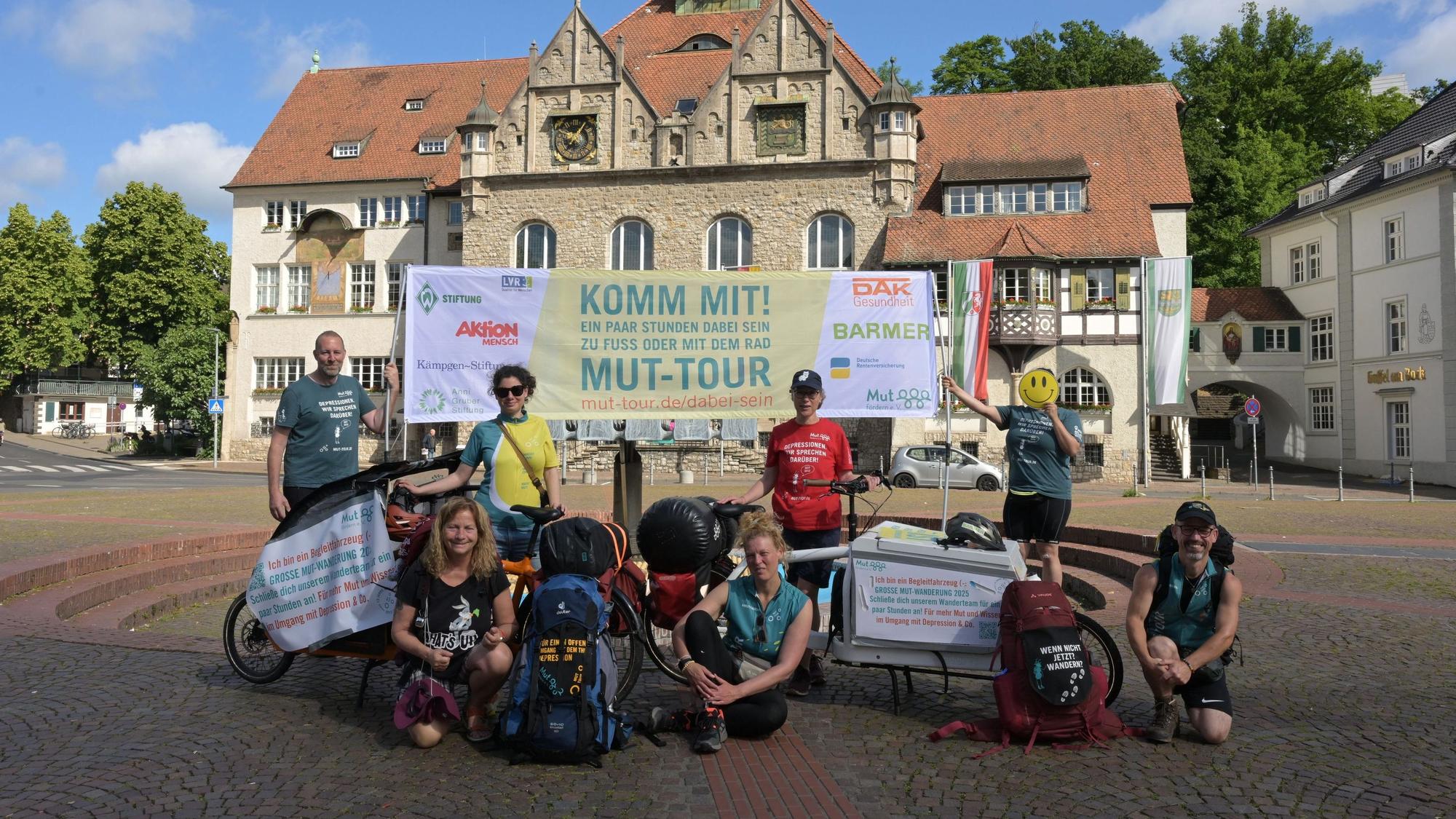 Sieben Wandernde der Mut-Tour stehen vor dem Papierbrunnen auf dem Konrad-Adenauer-Platz.
