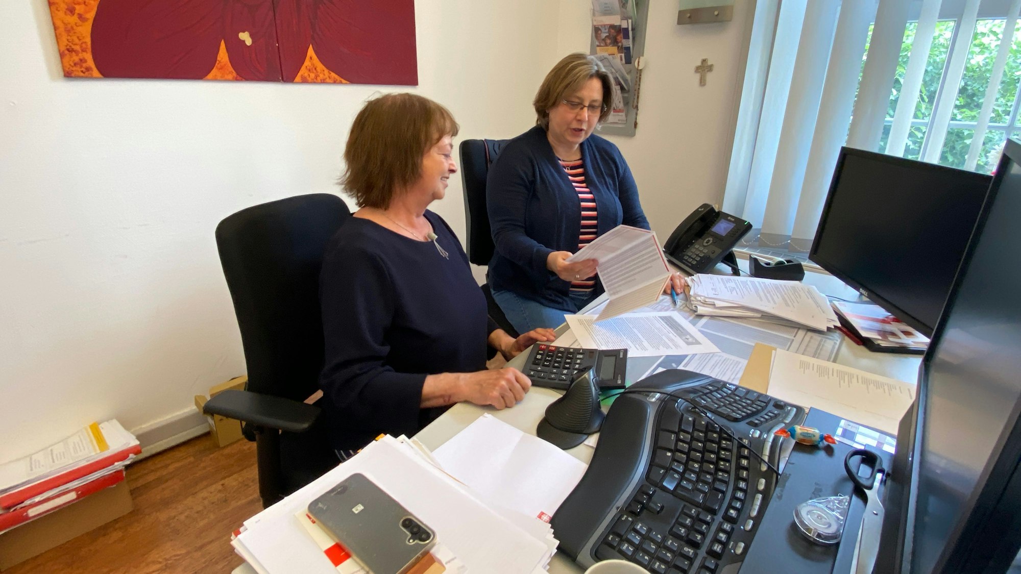 Zwei Frauen sitzen nebeneinander an einem Schreibtisch und schauen sich Unterlagen an.