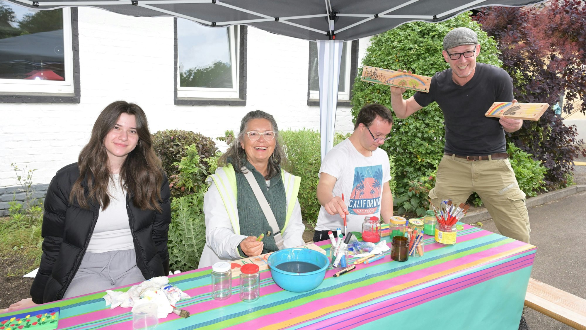 Drei Menschen sitzen vor einem Tisch und bemalen Schilder, einer steht daneben und hält die Schilder hoch.