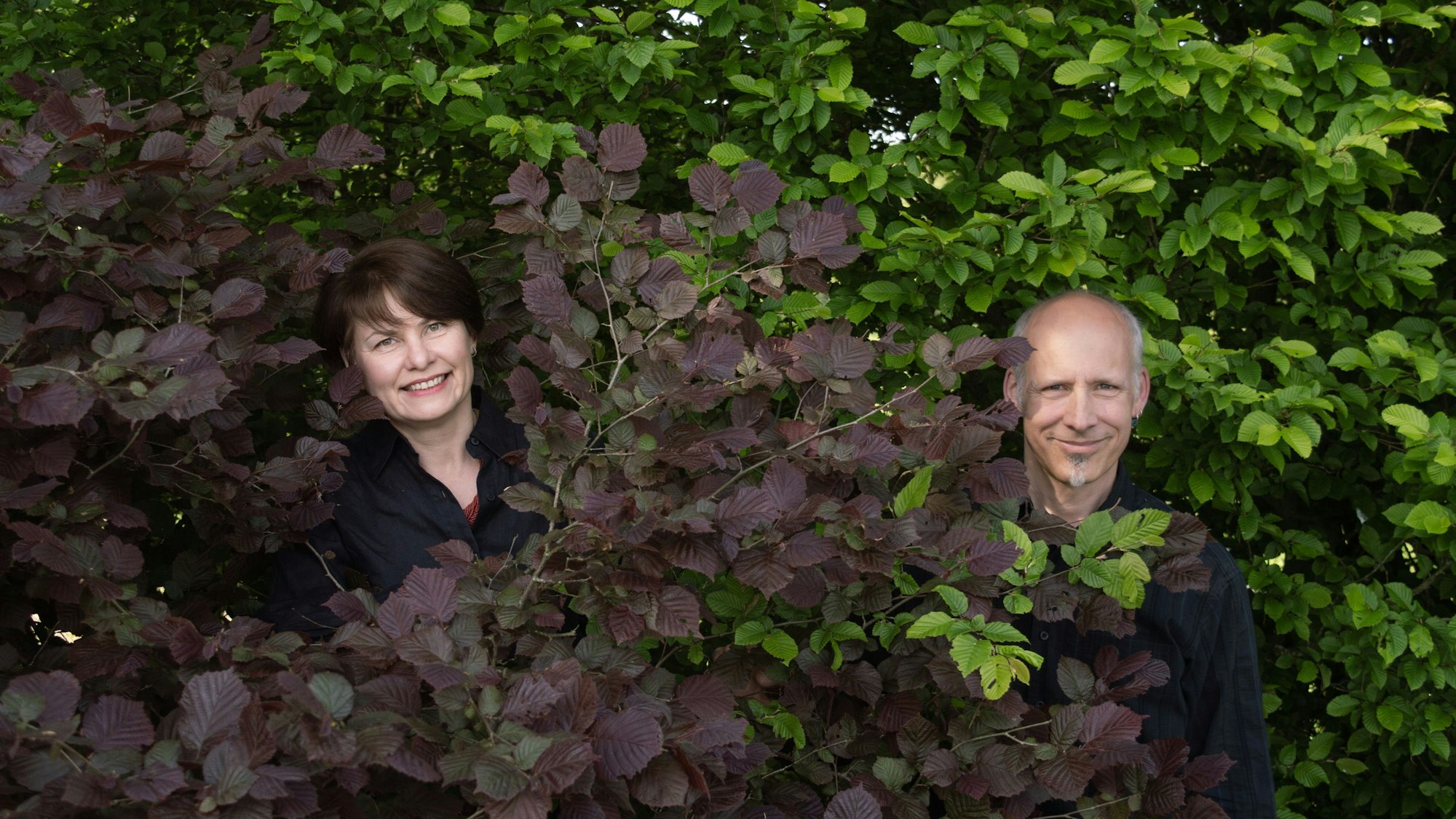 Irén Lill und Holger Faust-Peters schauen durch Zweige in die Kamera.