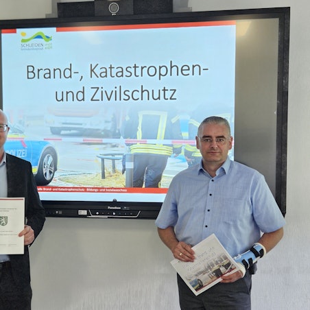 Ingo Pfennings (l.) und Wolfgang Fuchs stehen vor einem Bildschirm mit einer Fotocollage zum Brand-, Katastrophen- und Zivilschutz.
