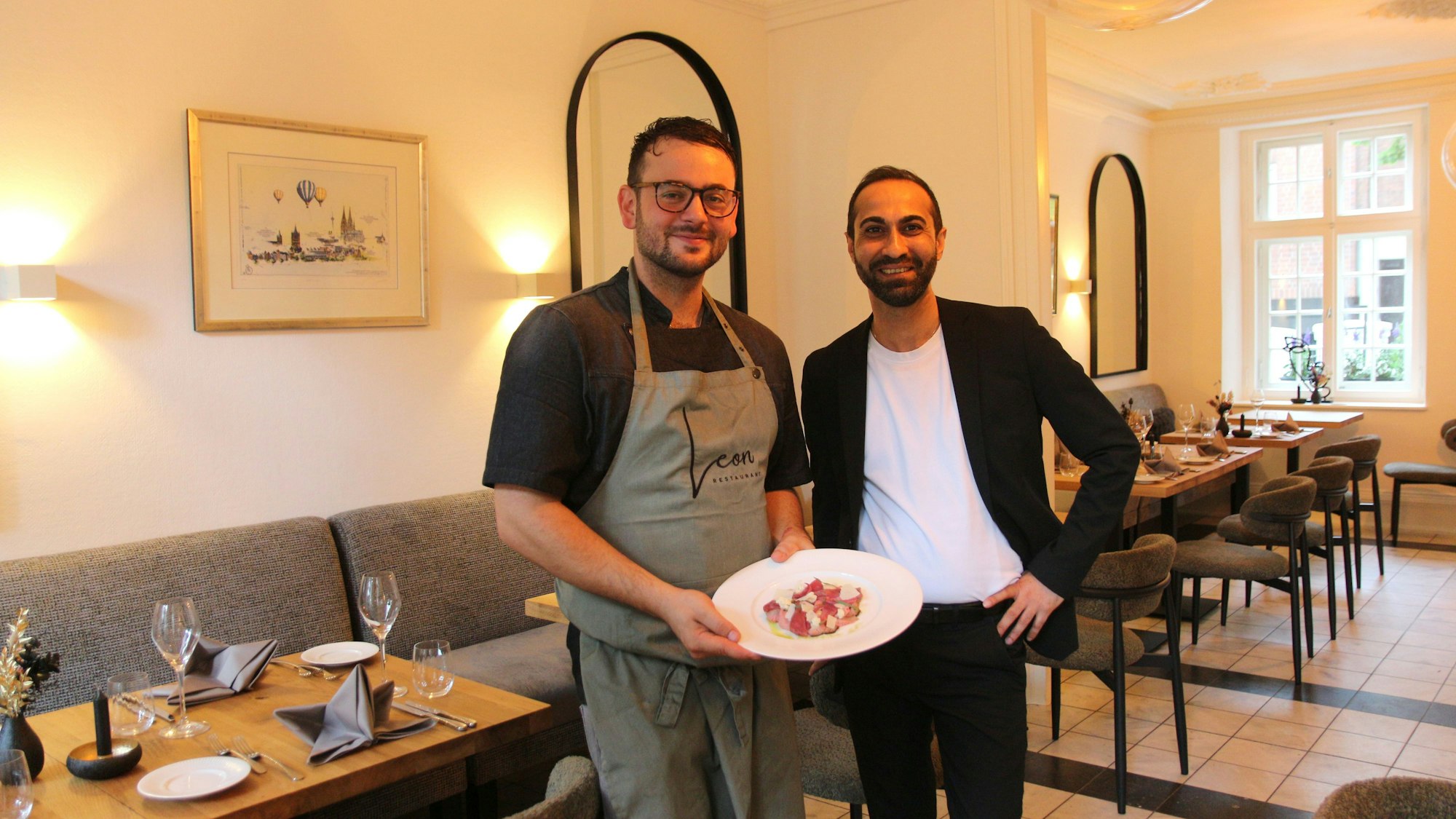 Zwei Männer lächeln in die Kamera, einer hält einen Teller mit einem Gericht in der Hand.