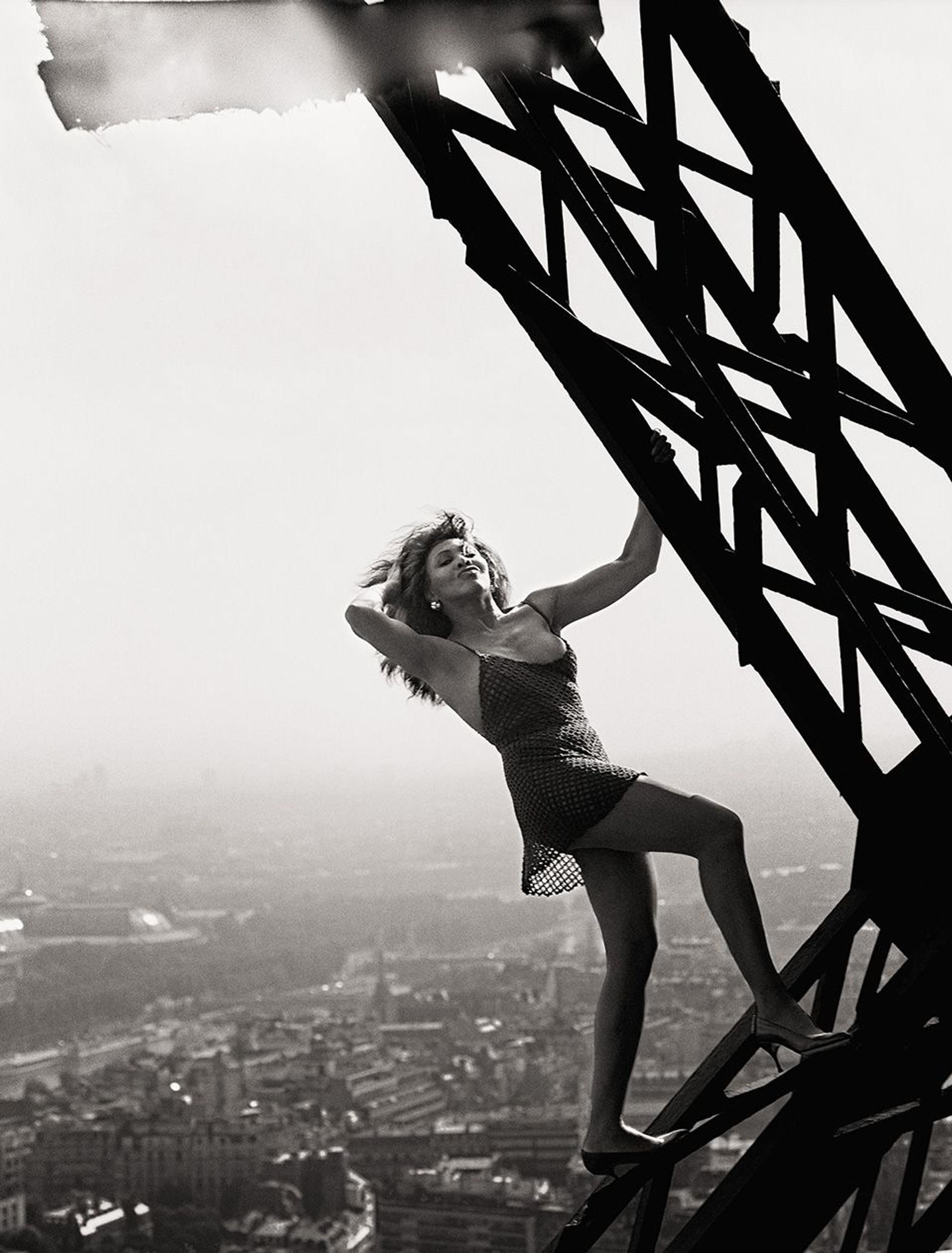 Tina Turner auf dem Eiffelturm. Mit der linken Hand hält sie sich am Fachwerk des Turms fest, mit der rechten streicht sie sich die Haare zurück. Ihr Blick verrät eigensinnigen Genuss. Sie trägt nur ein knappes Netzkleid, balanciert mit Stöckelschuhen auf dem schmiedeeisernen Träger.