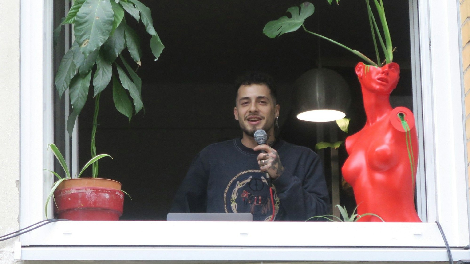 Ein junger Mann hält ein Mikrofon, er steht an einem geöffneten Fenster, links und rechts stehen Topfpflanzen.
