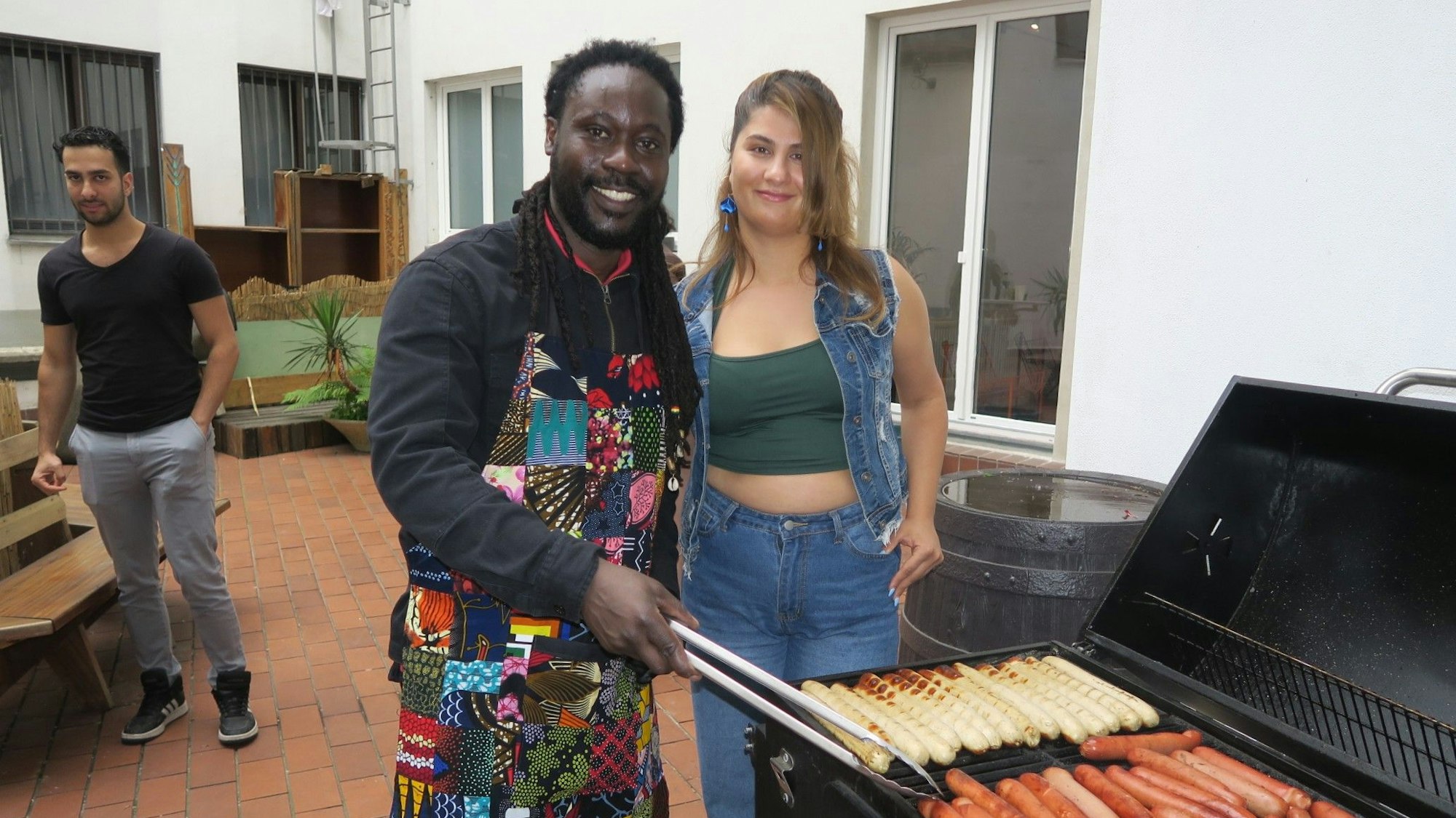 Ein dunkelhäutiger Mann mit Bart und bunter Schürze und eine junge Frau mit langem Haar stehen neben einem Grill, auf dem zwei verschiedene Sorten Würste liegen.