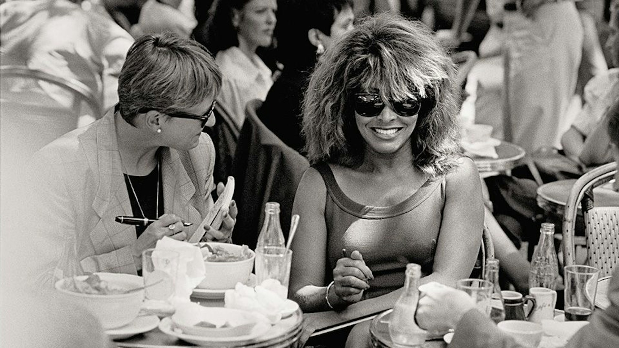 Tina Turner sitzt inmitten anderer Gäste in der Außengastronomie des Café de Flore. Sie trägt ein ärmelloses Top und eine Sonnenbrille.