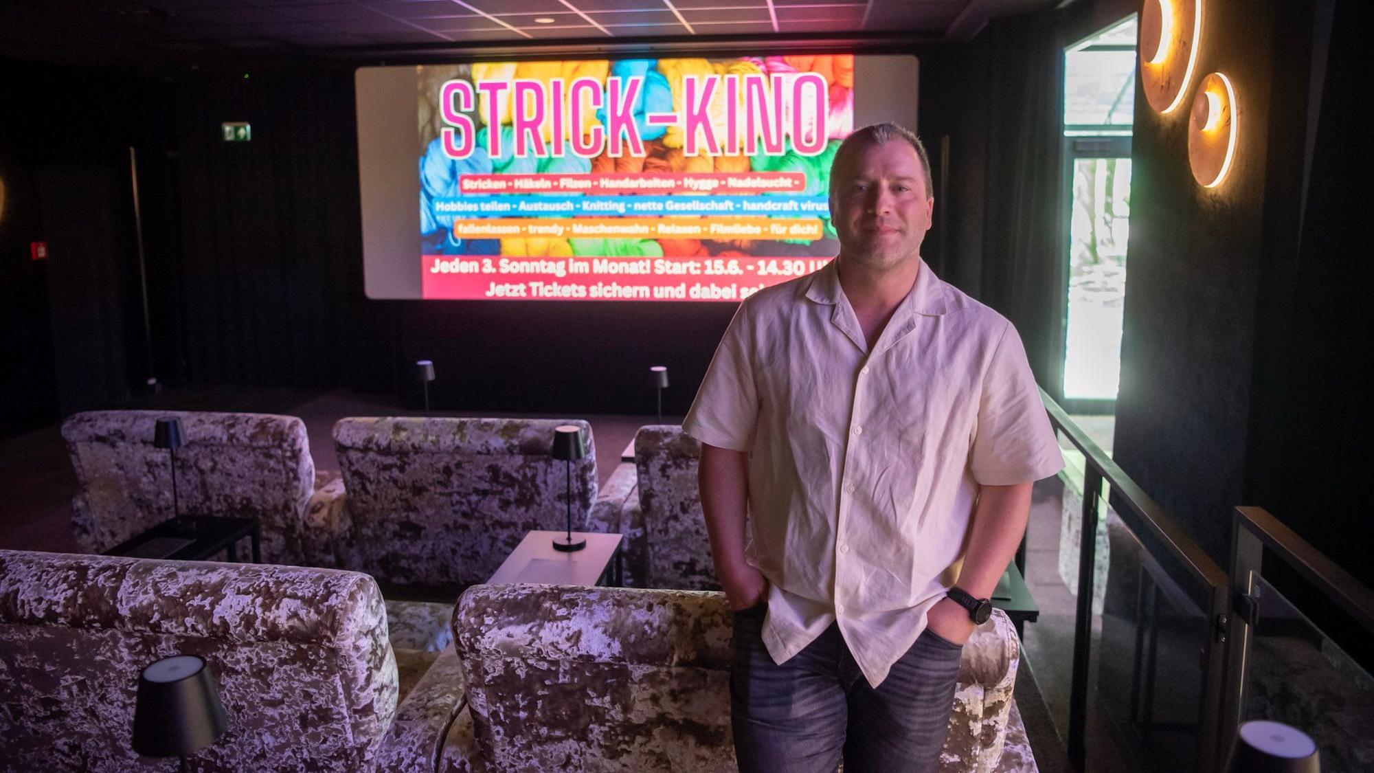 Sebastian Arenz steht an ein Plüschsofa gelehnt vor der Leinwand der Lichtspiellounge in Euskirchen, auf der in großen Buchstaben „Strick Kino“ steht.