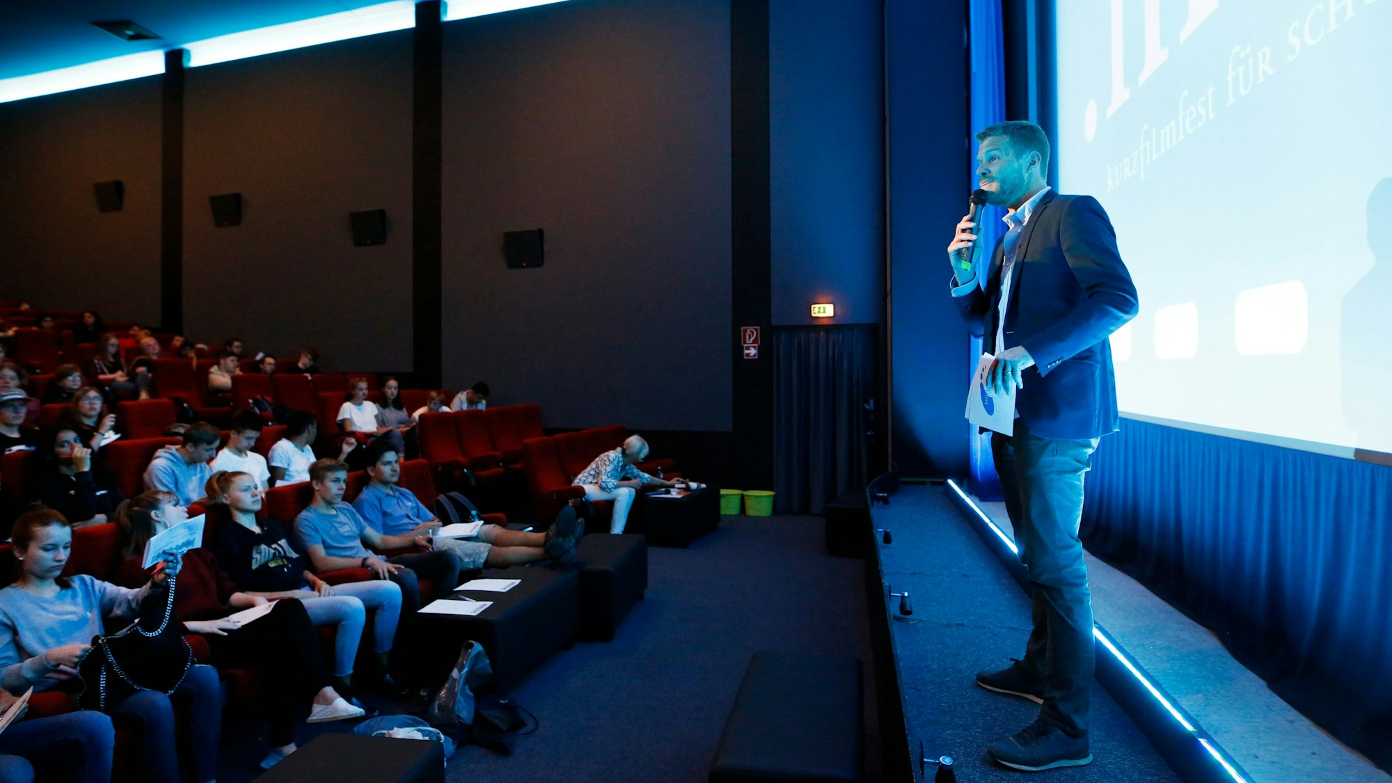 Beim Kurzfilmfest „.mov“ steht ein Moderator auf der Bühne des Filmforum NRW im Museum Ludwig in Köln. Schülerinnen und Schüler sitzen im Publikum.