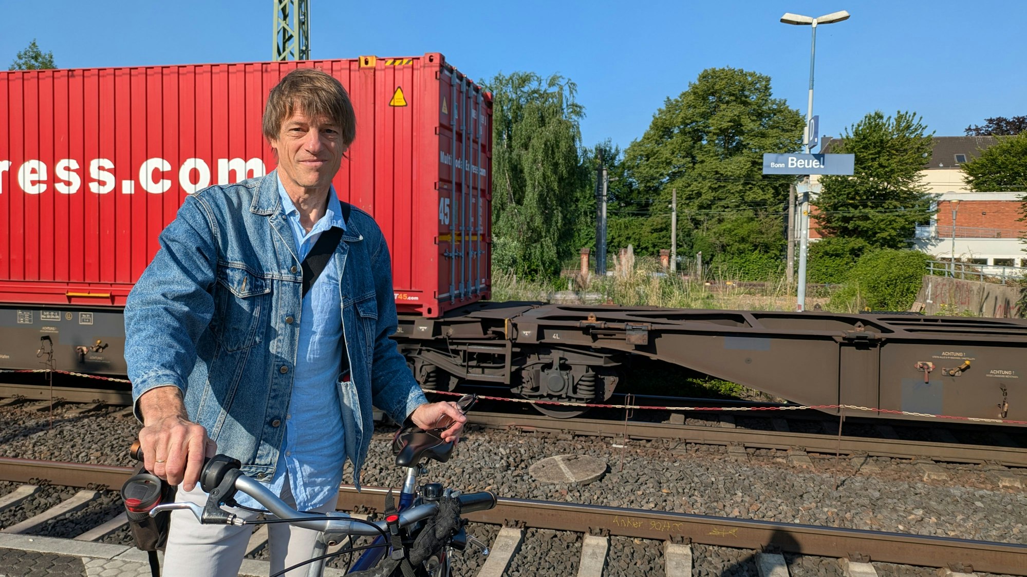 Ein Mann steht mit einem Fahrrad auf einem Bahnhof, im Hintergrund ein Güterzug.