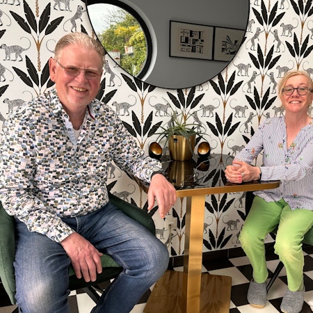 Ein Mann und eine Frau sitzen an einem kleinen Tisch vor einer gemusterten Tapete.