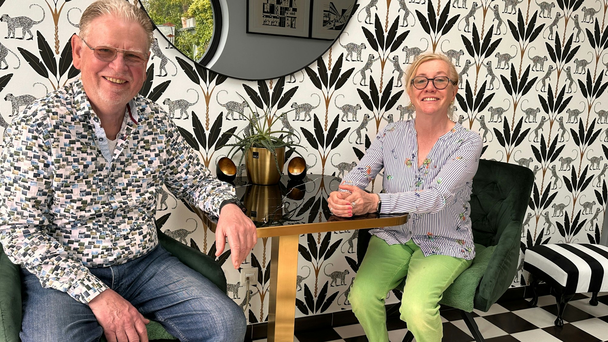 Ein Mann und eine Frau sitzen an einem kleinen Tisch vor einer gemusterten Tapete.