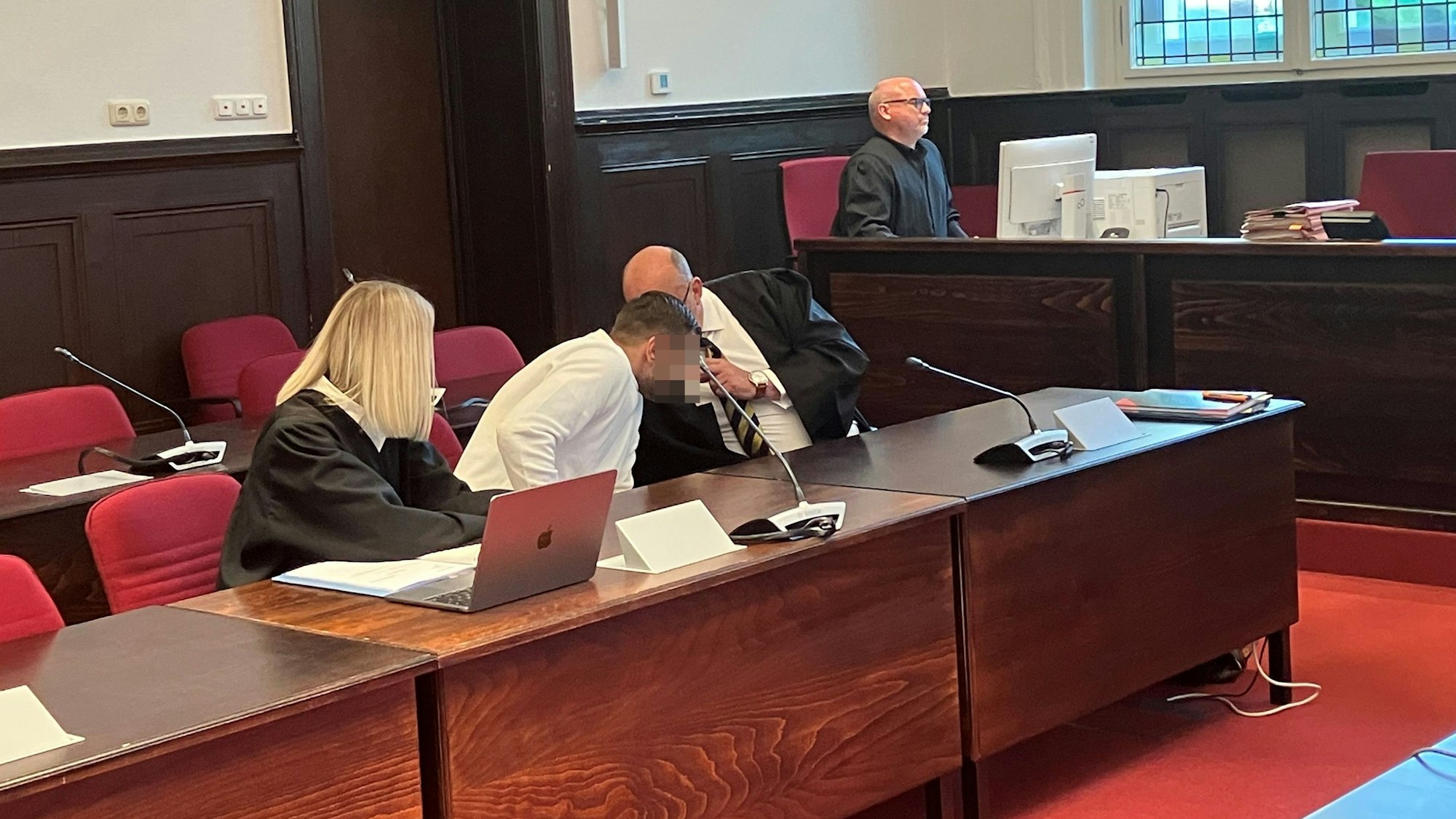 Ein Mann in weißem Oberteil sitzt zwischen zwei in schwarzen Roben gekleideten Anwälten hinter einem dunkelbraunen Holztisch.