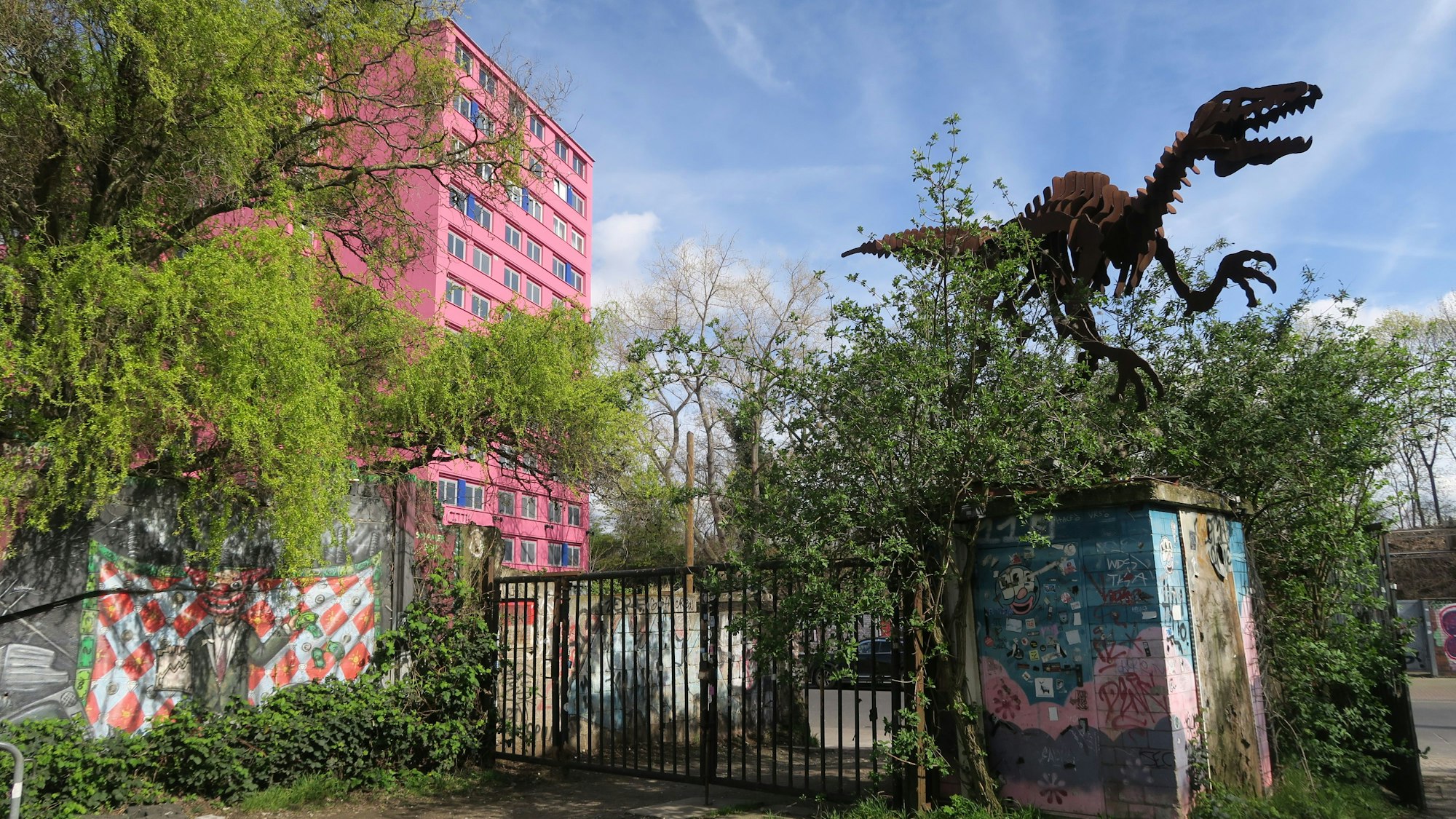 Hinter einem rostigen, halb zugewucherten Tor steht eine riesige Dinosaurier-Skulptur, auch ein mehrstöckiges pinkes Gebäude ist zu sehen.