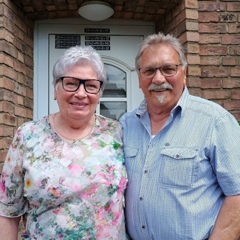 Klara Bloch trägt eine Bluse mit Blumen, ihr Mann ein hellblaues Hemd. Sie stehen vor ihrem Haus und lächeln in die Kamera.