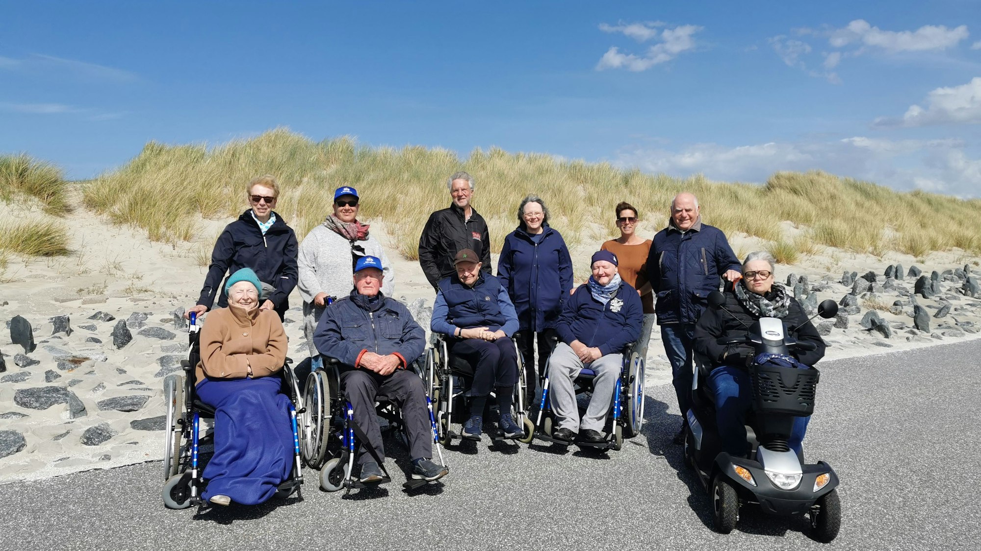 Fünf Menschen im Rollstuhl sowie Pflegende und Angehörige auf Norderney vor den Dünen.
