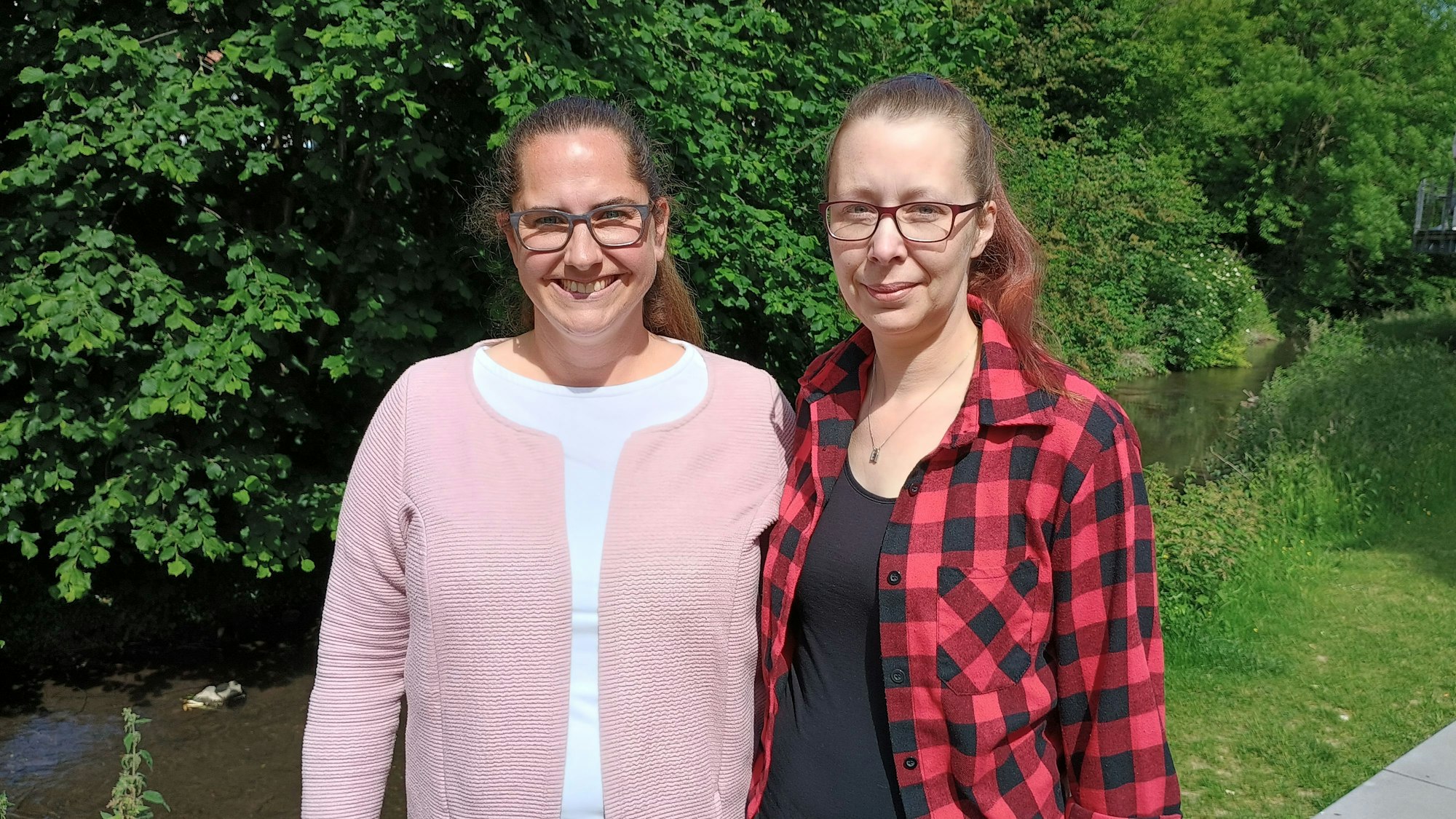 Die beiden stehen beieinander und lächeln. Hinter ihnen fließt die Kleine Erft, am Ufer dichtes Blattwerk der Bäume.