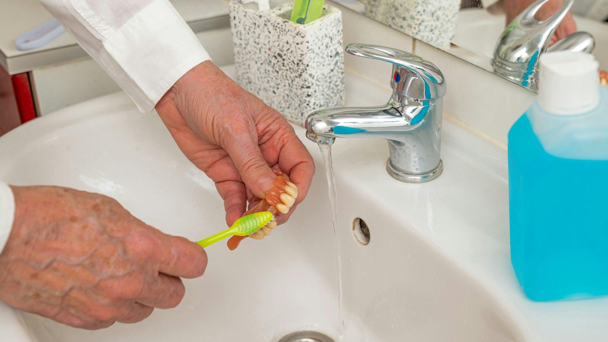 Hände eines Mannes, der sein Gebiss mit einer Zahnbürste im Waschbecken unter dem Wasserhahn reinigt.