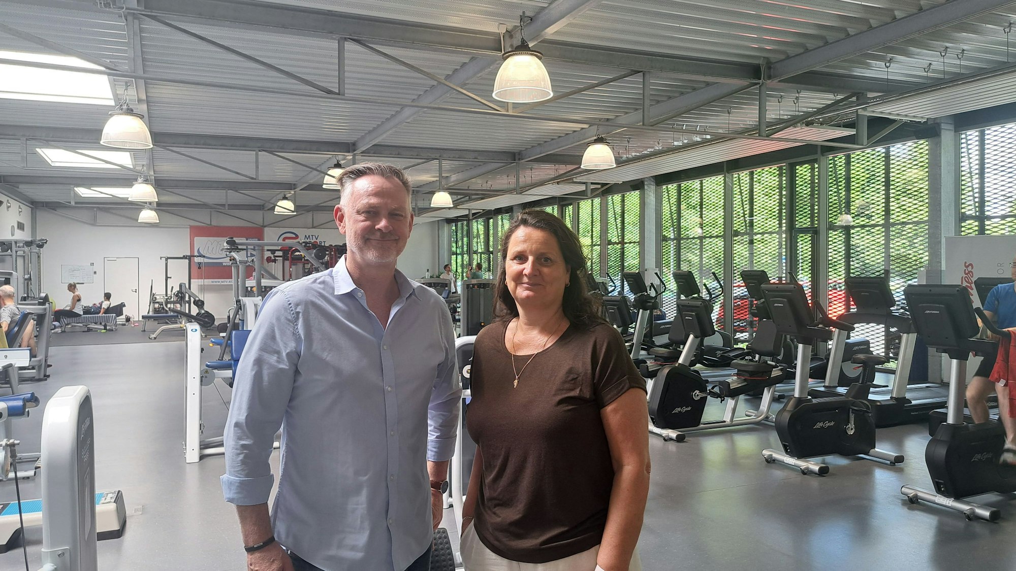 Ein Mann in einem hellblauen Hemd und eine Frau in einem braunen Shirt stehen in einem Fitnessstudio.