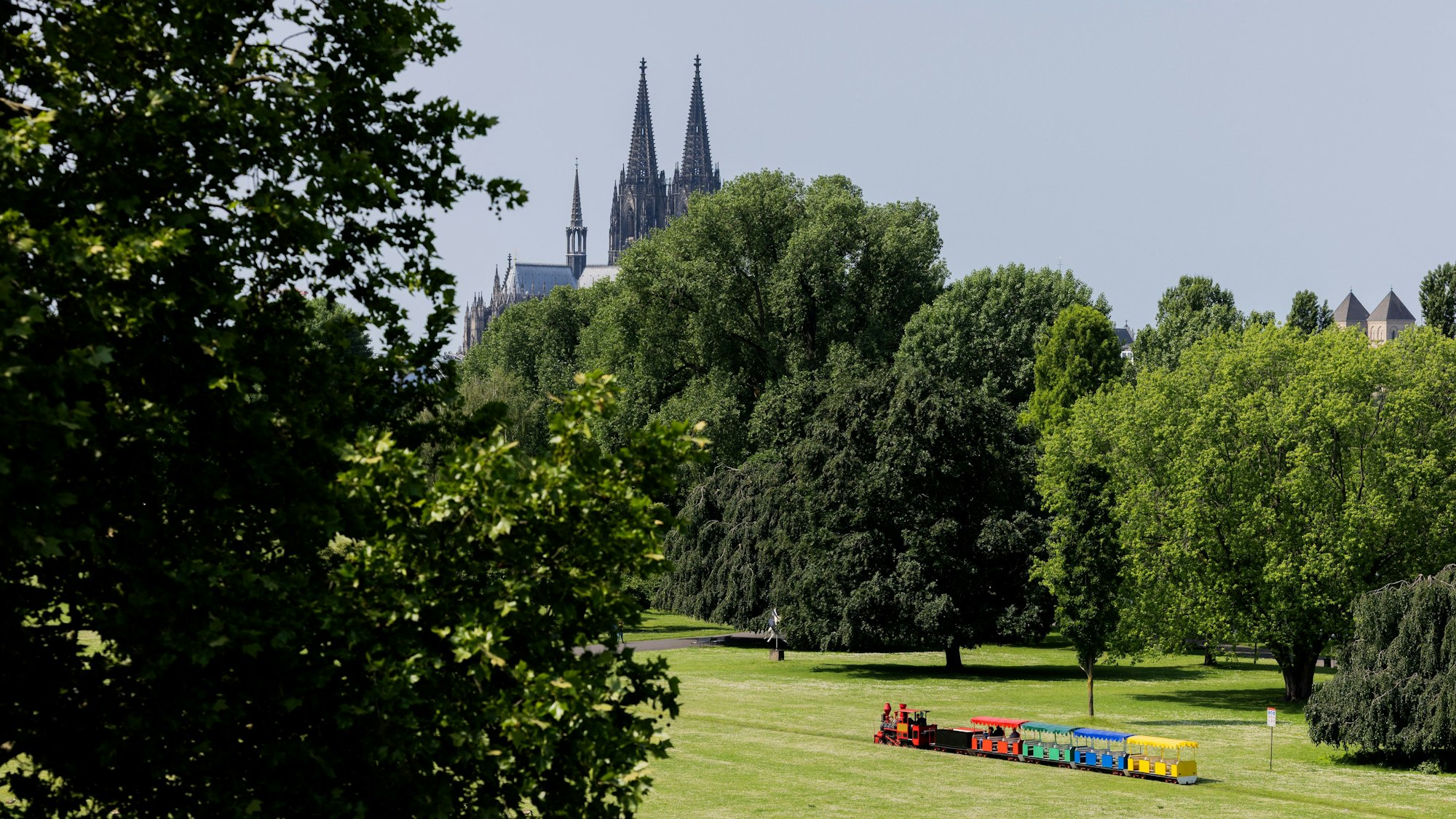 02.06.2025, Nordrhein-Westfalen, Köln: Eine Kleinbahn fährt bei Sonnenschein vor der Kulisse des Kölner Doms durch das Grün im Rheinpark. Foto: Rolf Vennenbernd/dpa +++ dpa-Bildfunk +++