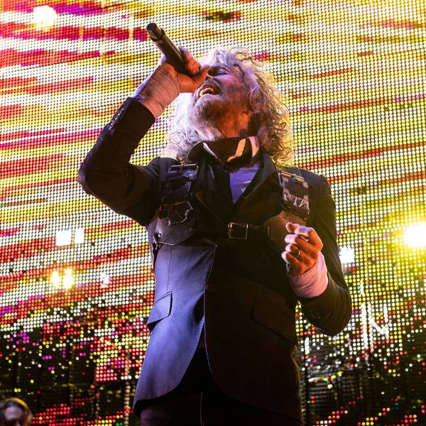 Wayne Coyne singt mit angewinkelten Arm und zurückgeworfenen Kopf ins Mikro. Er trägt einen dunkelblauen Anzug vor gelb-roter Videoleinwand.